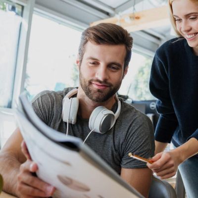 Two coworkers collaborate at a desk: man with headphones reads a document while a woman points with a pencil.