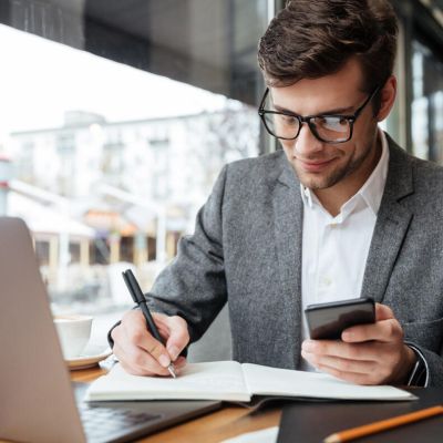 Man in a gray blazer writes in a notebook while glancing at his smartphone at a cafe table with a laptop nearby.